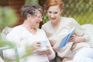 A senior woman with a mug of coffee next to a caregiver with a book