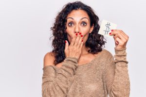 A middle aged Hispanic woman looking shocked holding up a $ sign as an indication of how caregiving and finances can be complex