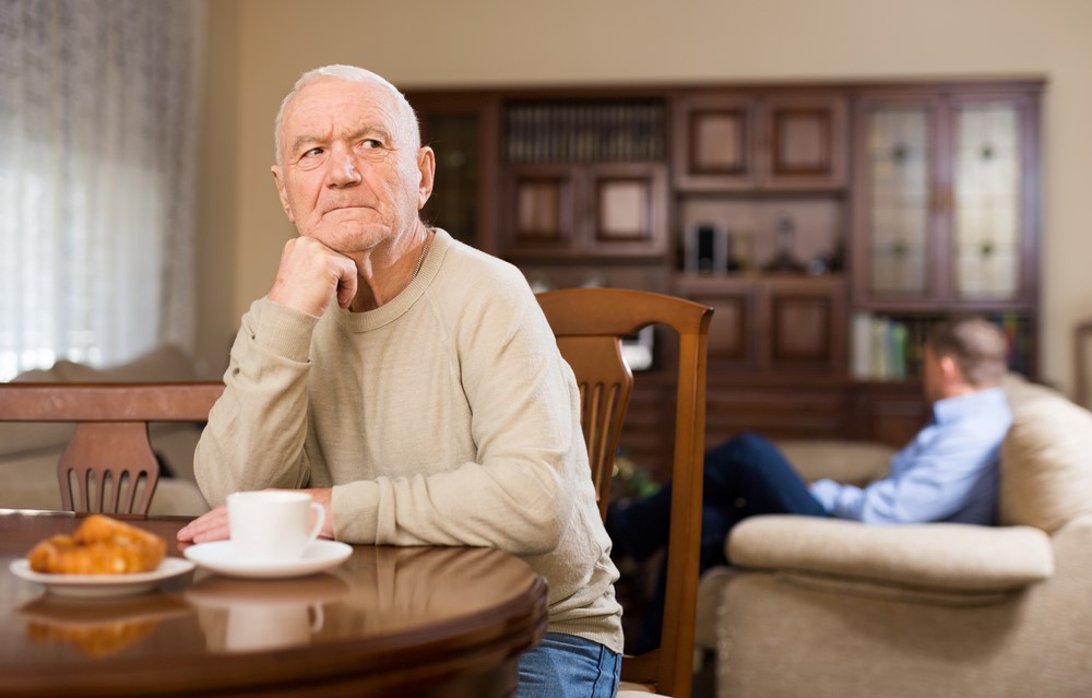 An upset elderly man sitting alone at a table, with his son in the background