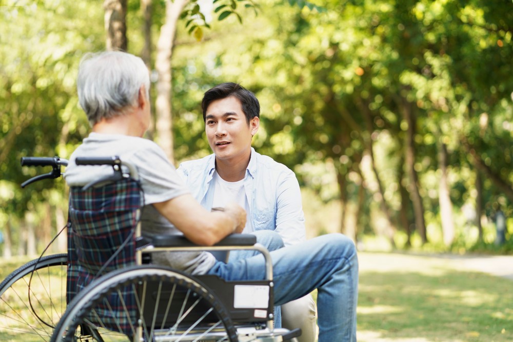 An Asian senior sitting in a wheelchair outside, talking to his son