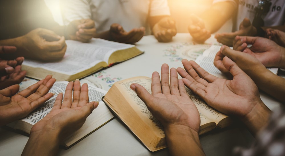 A group of people sitting around a table praying with their palms open over bibles