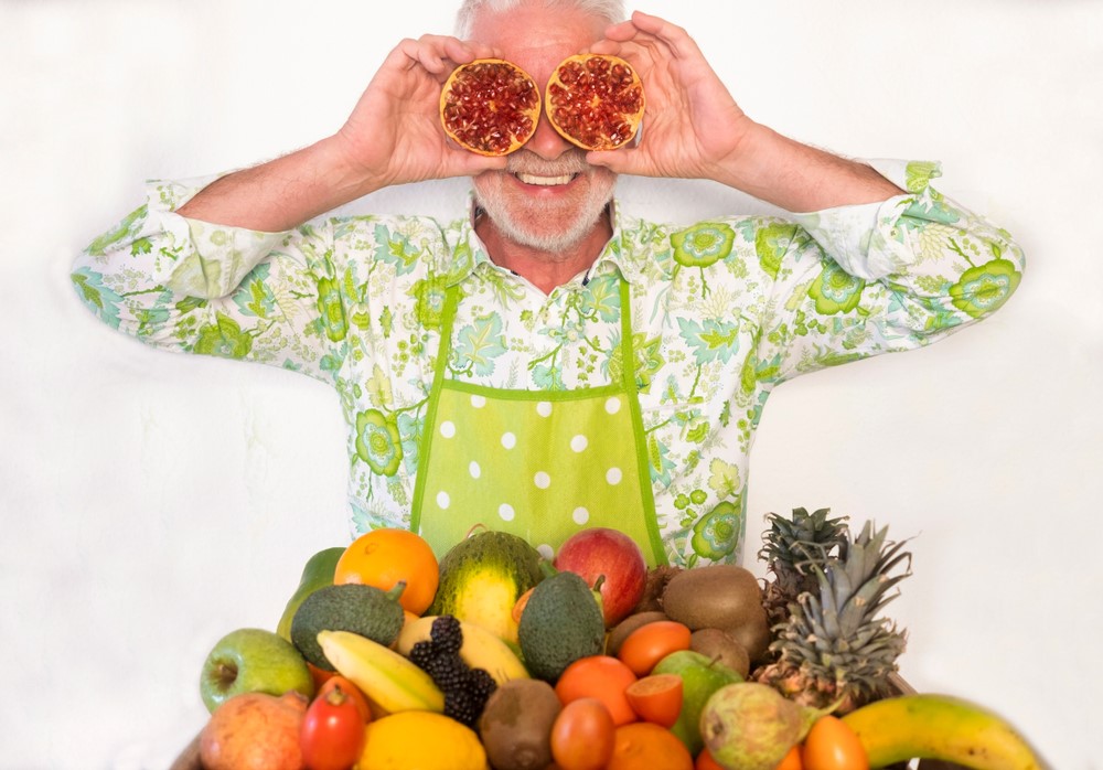 A senior man holding pomegrantes to his eyes with a stack of fruit below him