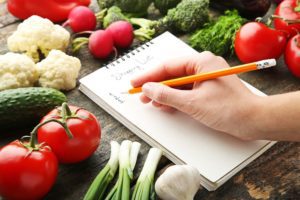 Someone writing a grocery list while surrounded by tomatoes, cauliflower and other fresh vegetables. highlighting the idea of healthy recipes for seniors