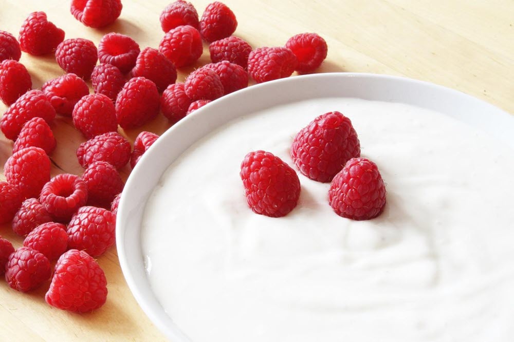 A white bowl of yogurt with three raspberries on top, next to more raspberries