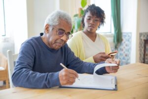 A senior man writing on paper while a younger woman looks on, highlighting the idea of becoming a home care nurse or a caregiver