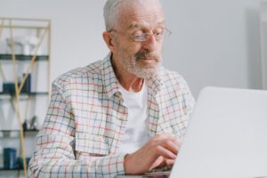 An older man smiling while he is looking at a laptop, doing an online course