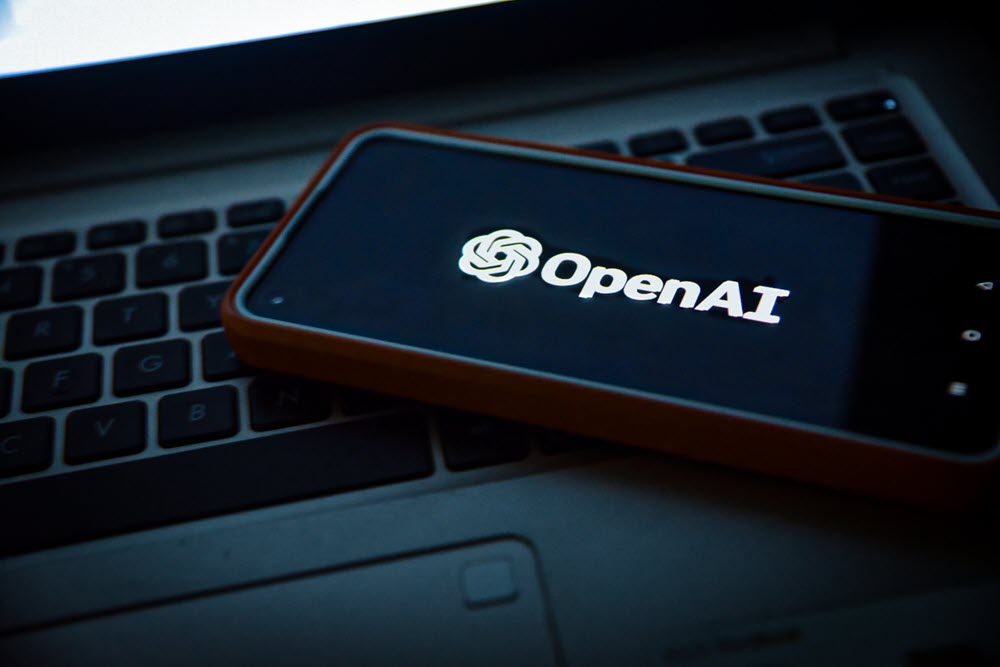 A laptop with a phone on top that is showing the OpenAI logo